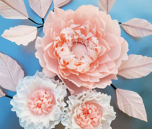 Handmade paper floral arrangement featuring a prominent blush pink peony with ruffled petals and three smaller white blossoms with textured centers, all set against a pastel blue background emphasizing the artistry and three-dimensional aspect of the paper