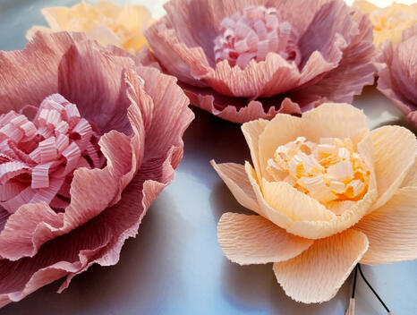 serene display of three large mauve paper flowers with detailed pink centers and two smaller peach blooms, complemented by delicate white leaves on a soft blue background.